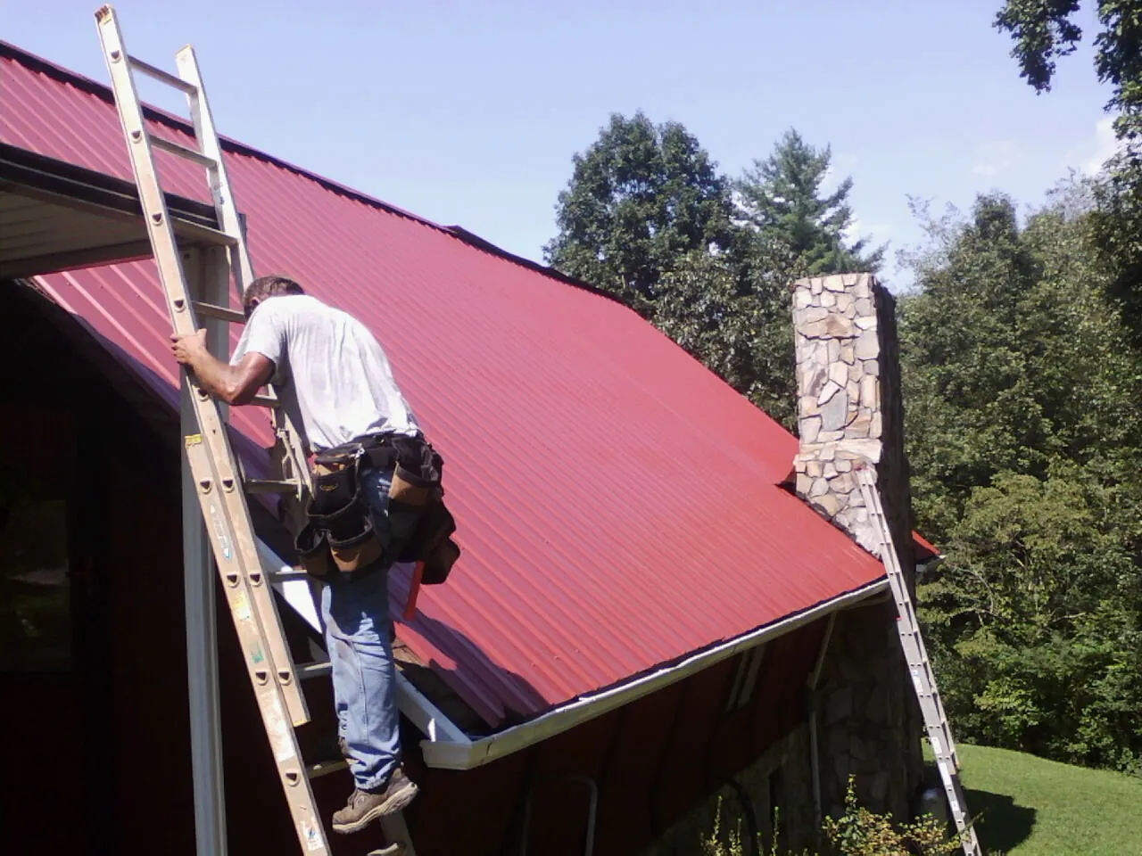 Roofer on a ladder beside a red metal roof and stone chimney — Soffit & Fascia Repair by Roofing Brothers in Ferguson